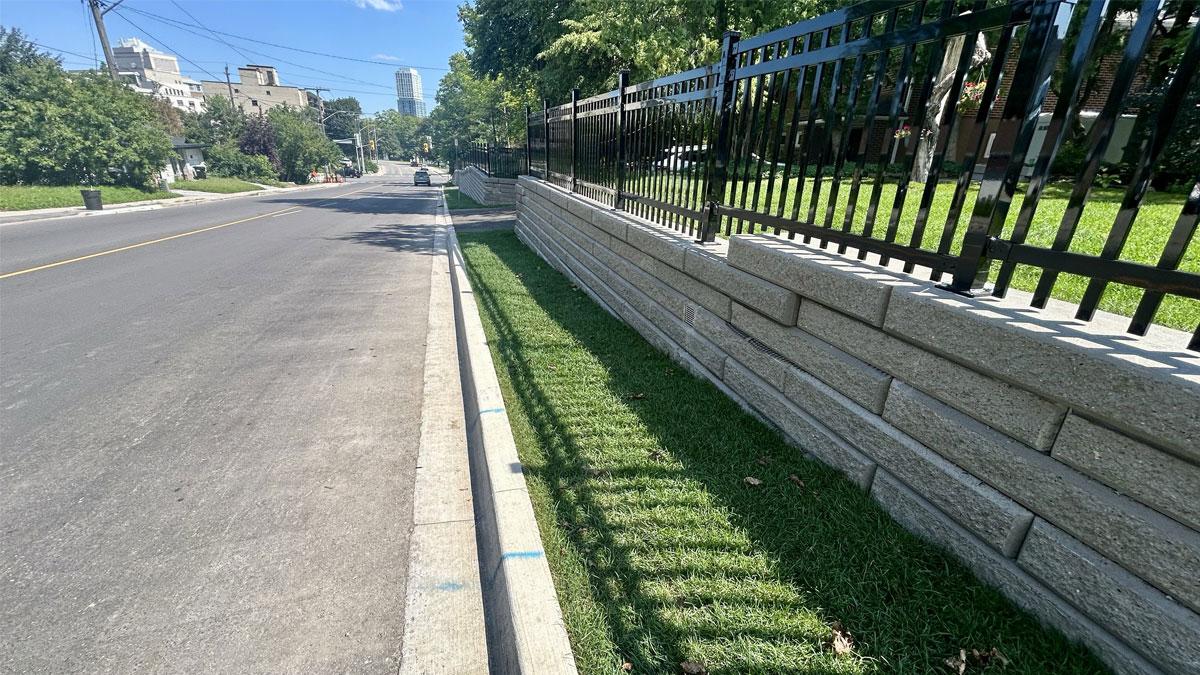 A sidewalk along a street with green grass and a stone wall beside a fence.