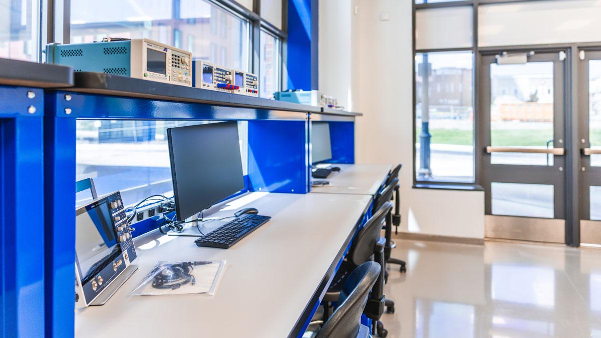 Bright, modern workspace featuring multiple computers and blue shelving. Large windows visible.