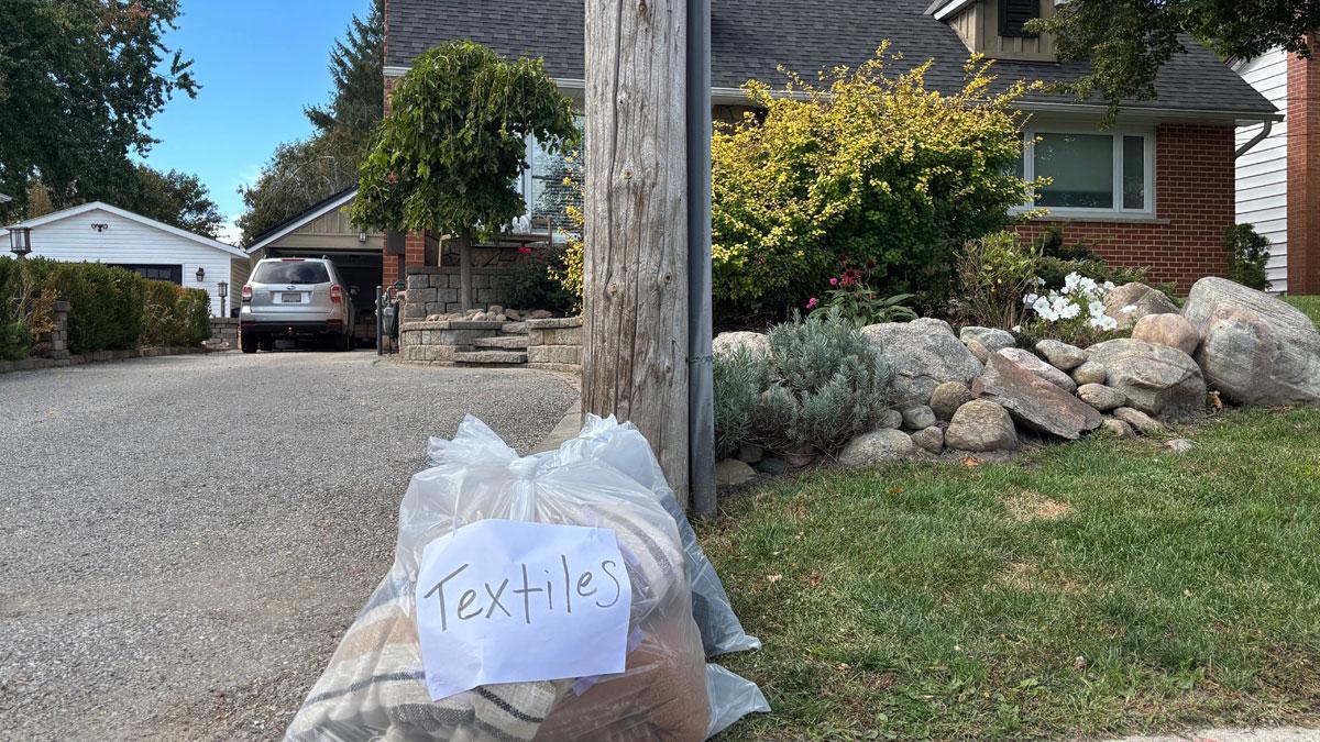 A clear bag labeled "Textiles" sits on the grass by the curb, with a house and car in the background.