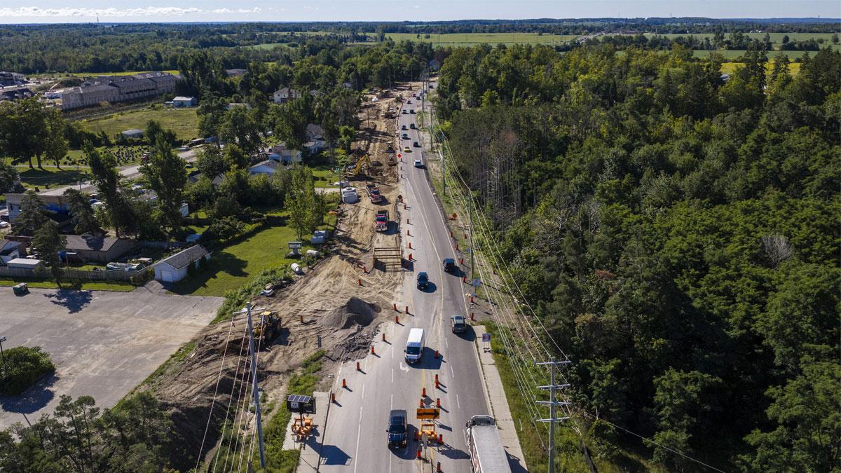 Construction scene along a road with heavy machinery and vehicles among trees and fields.
