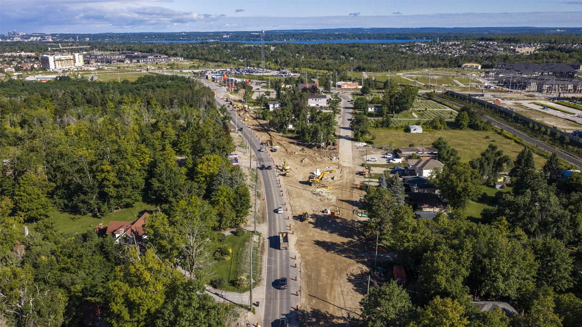 Aerial view of a road with construction, surrounded by greenery and distant buildings.