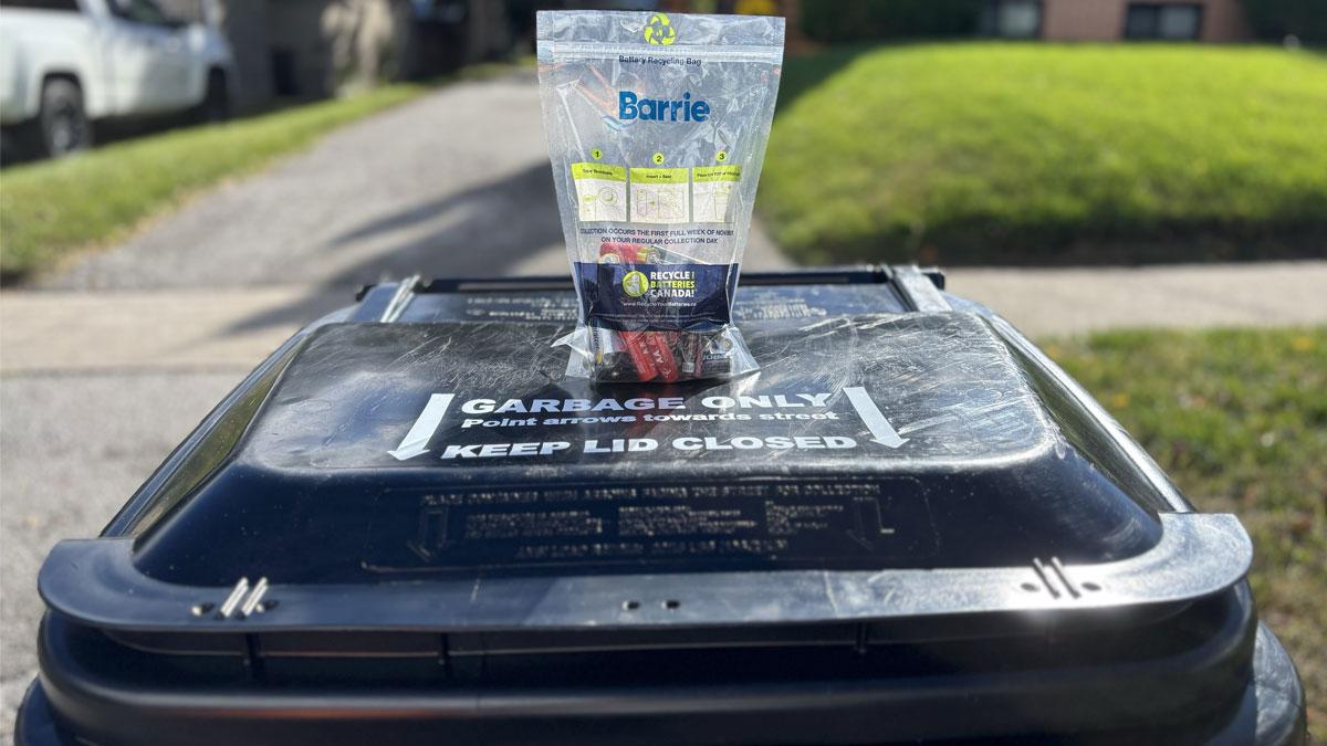 A clear bag with the Barrie logo filled with batteries on a black garbage cart outside, with grass in the background.