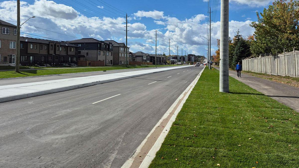 A wide, empty street with a centre median, bordered by grassy areas and residential buildings.