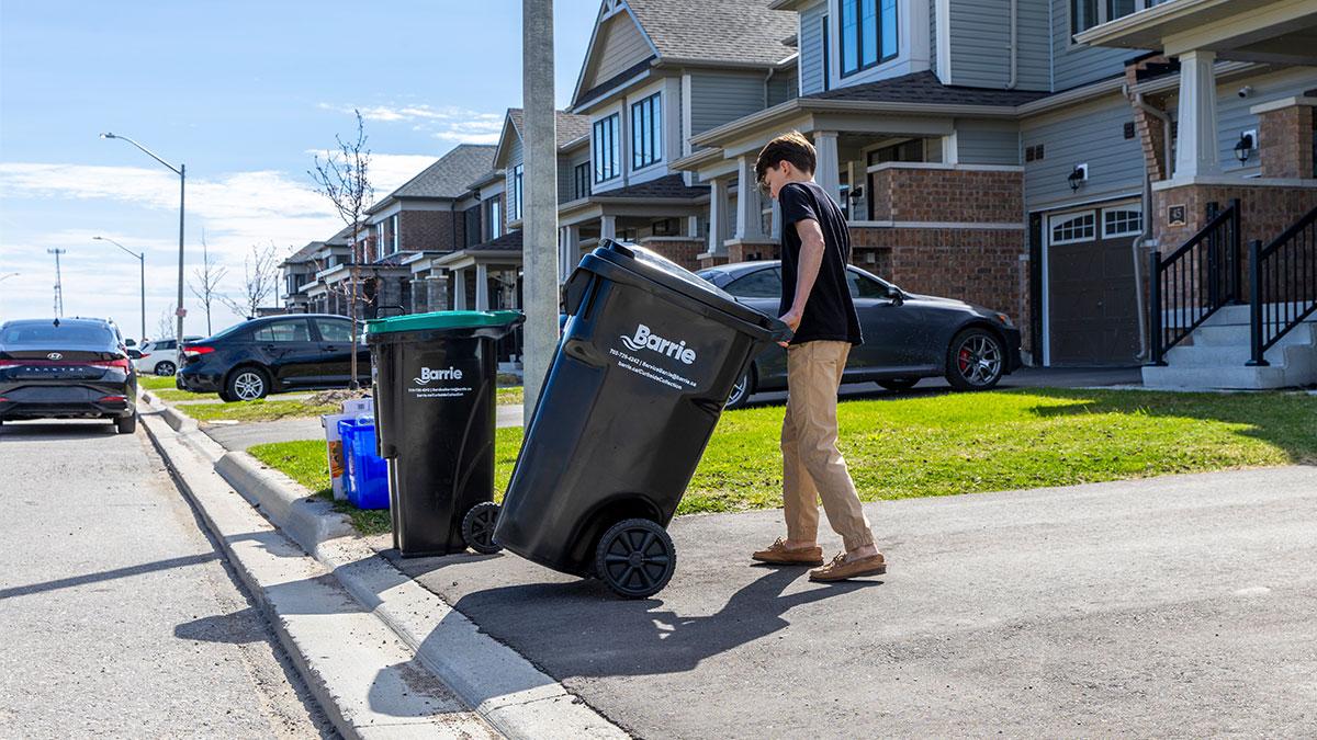 Person rolling a Barrie-branded garbage cart on a residential street lined with townhouses on a sunny day.