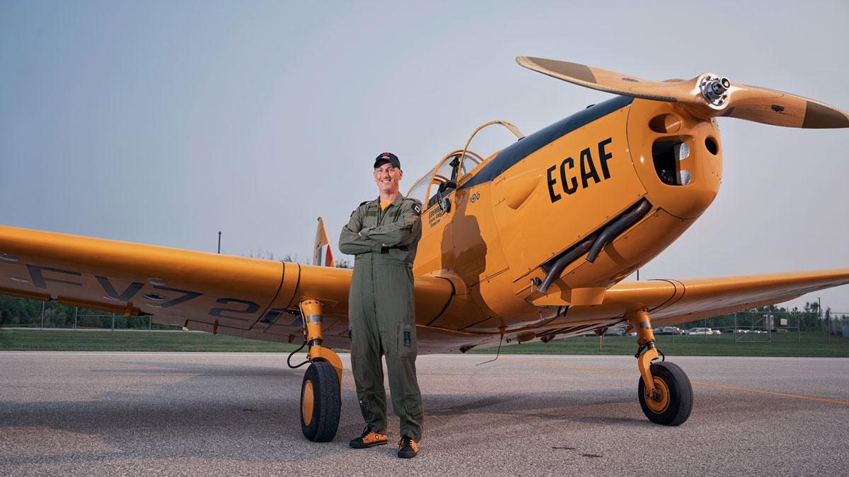 Pilot in green jumpsuit stands in front of a yellow vintage aircraft on a runway.