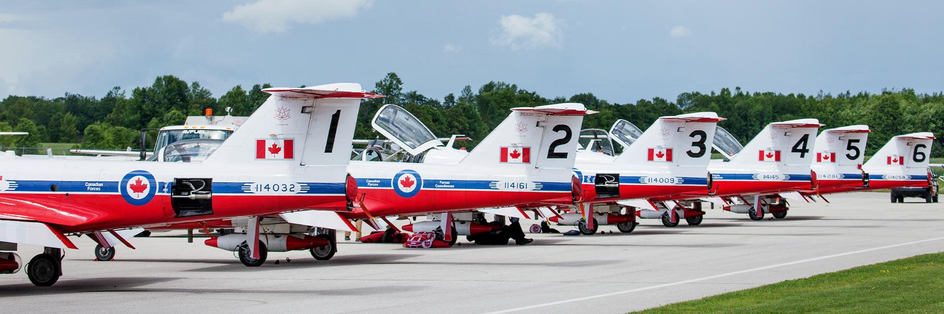 Row of Canadian jet aircraft parked on tarmac with blue sky and trees in background.