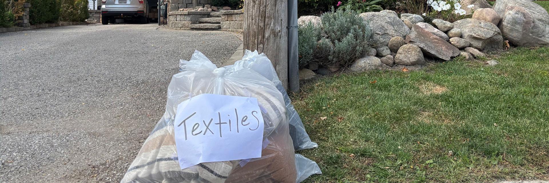 A clear bag labeled "Textiles" sits on the ground at the end of a driveway.