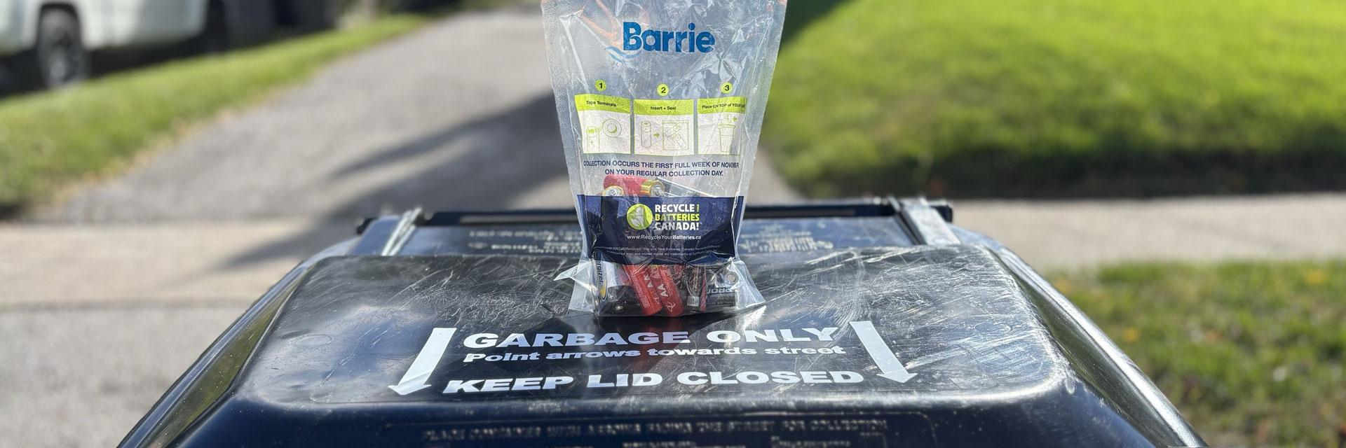 A clear bag with the Barrie logo filled with batteries on a black garbage cart outside, with grass in the background.