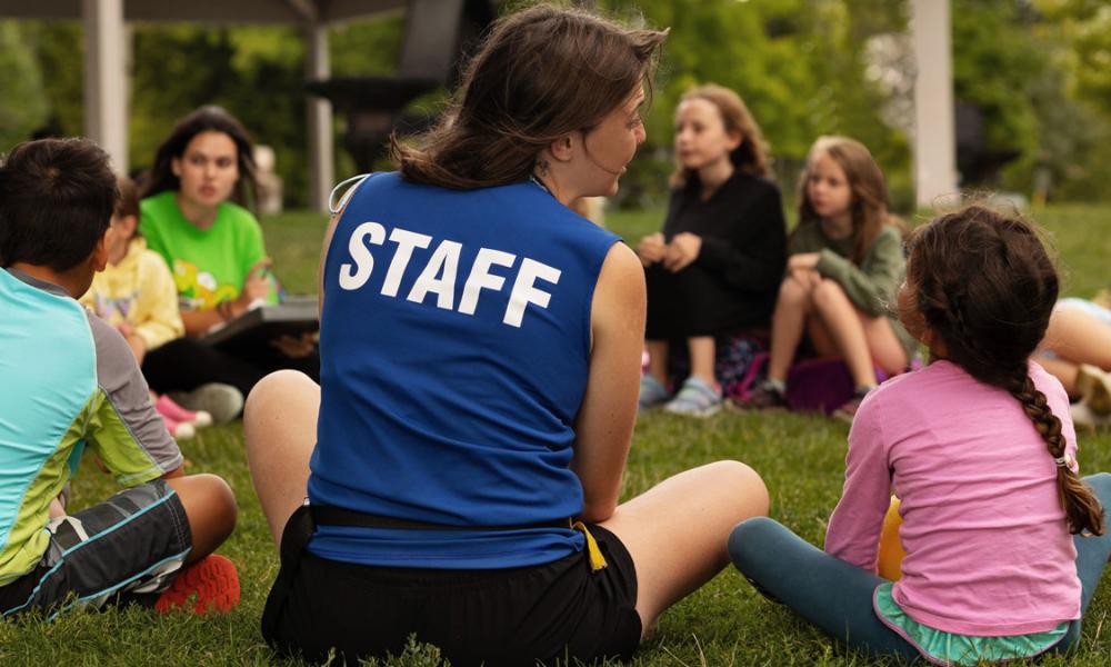 Woman sitting on grass with children, facing away from camera, with "STAFF" on back of shirt