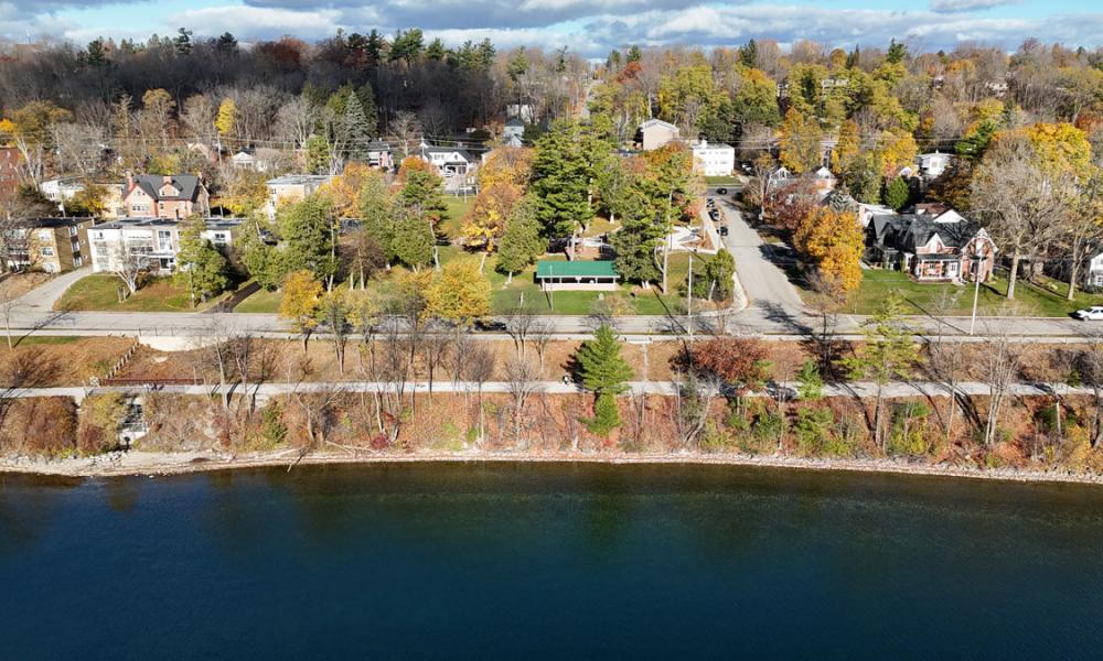 Aerial view of a trail and road beside a body of water, featuring residential buildings surrounded by trees with autumn foliage.
