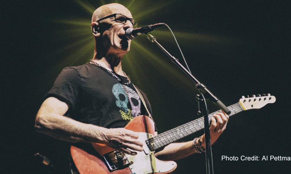 Kim Mitchell playing an electric guitar and singing on stage, with a bright spotlight behind him.
