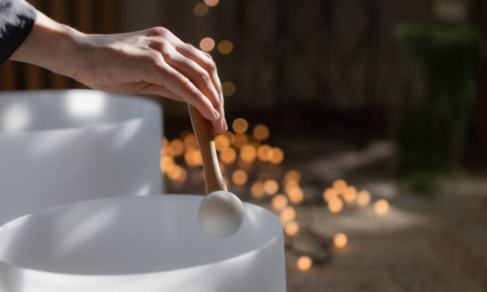 A hand holds a mallet above a white crystal singing bowl, with warm lights in the background.
