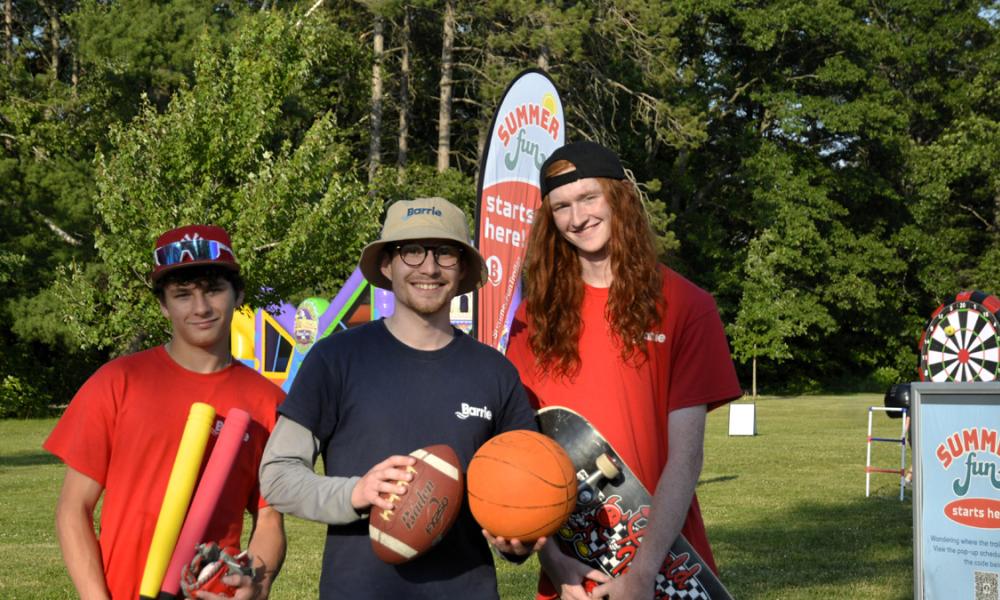 Three young men stand outdoors, holding sports equipment with trees and colorful banners in the background.