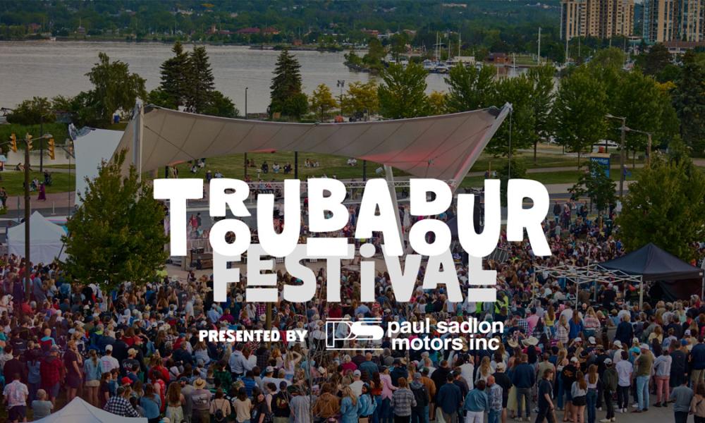 Crowd enjoying an outdoor festival near a lake, with trees and buildings in the background. Troubadour Festival Presented by Paul Sadlon Motors.