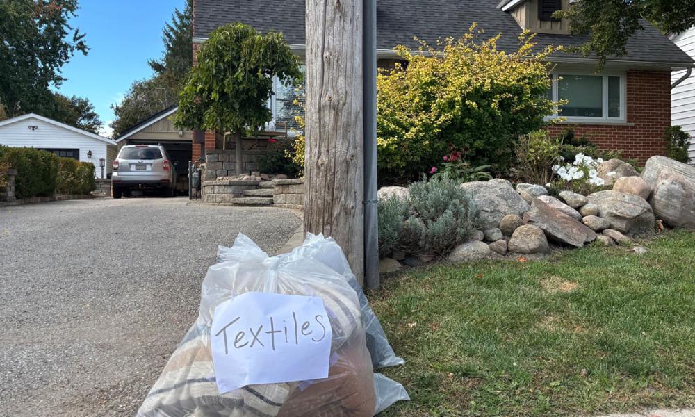 A clear bag labeled "Textiles" sits on the grass by the curb, with a house and car in the background.