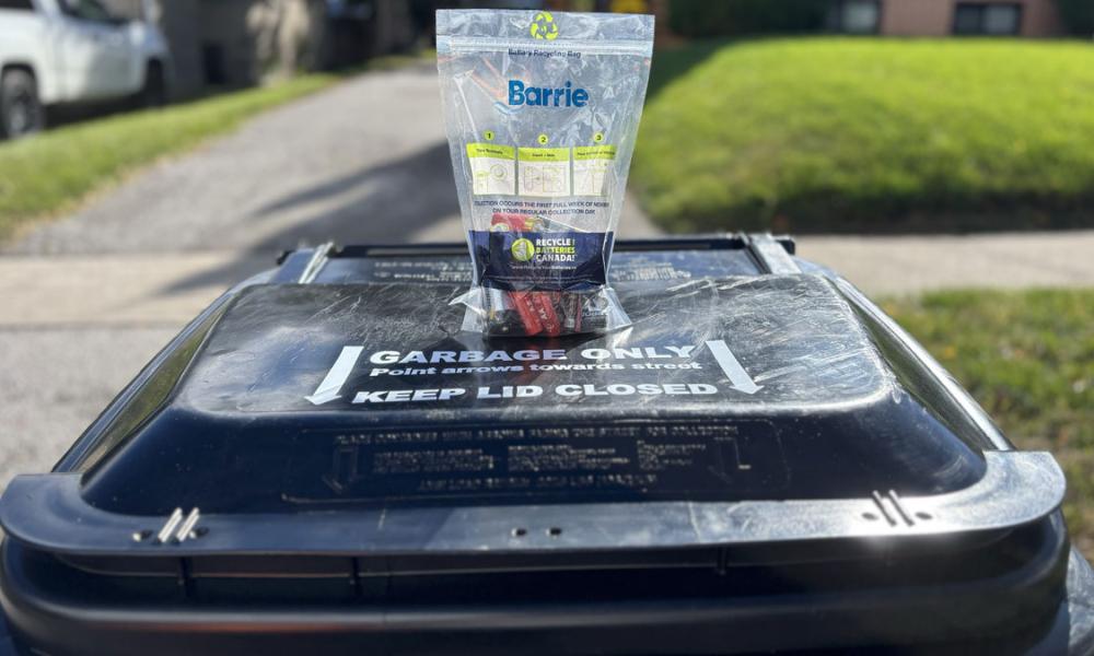 A clear bag with the Barrie logo filled with batteries on a black garbage cart outside, with grass in the background.